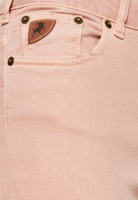 Vaaleanpunaiset farkut, joissa on sileä pinta, ja kultasävyiset yksityiskohdat. Takataskussa on ruskea nahkalaatta, jossa on härän siluetti.