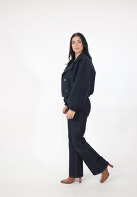 Granatowa krótka kurtka z dużymi klapami i złotymi guzikami, zestawiona z ciemnymi dzwonowatymi spodniami z denimu oraz brązowymi klapkami na obcasie. Gładka tekstura tkaniny.