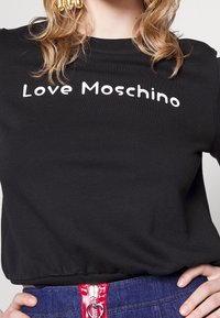 Černé tričko s nápisem "Love Moschino" bílým písmem. Měkká tkanina, kulatý výstřih a detaily na nařaseném lemu. Nosené s modrými džíny.