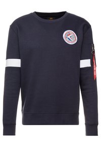 Sweatshirt marine à col rond, rayure blanche sur les manches et un patch représentant un astronaute ; poche zippée avec un détail de tag rouge.
