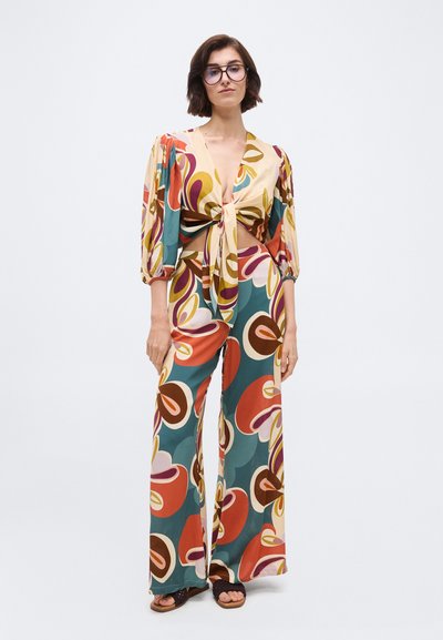 Γυναίκα που φοράει πολύχρωμο, φαρδύ παντελόνι με σχέδια και ταιριαστό μακρυμάνικο τοπ με δέσιμο μπροστά, σε συνδυασμό με μαύρα σανδάλια, στέκεται μπροστά σε γκρι απλό φόντο.