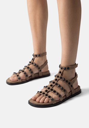 Pies con sandalias gladiador marrones con tachuelas redondas negras y tiras en el tobillo sobre un fondo claro.