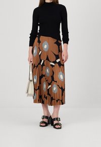 Donna in top nero a maniche lunghe, gonna a pieghe marrone con grande motivo floreale, sandali neri con tacco, e che tiene una borsa beige.