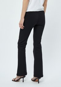 Pantaloni neri a zampa d'elefante con una texture liscia, dotati di vita media e tasche posteriori, abbinati a sandali neri con tacco alto e fascia.