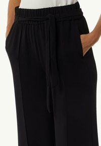Schwarze lockere Hosen mit breitem Bund, gebundenem Gürtel und Seitentaschen; glattes Gewebe mit weicher Textur.