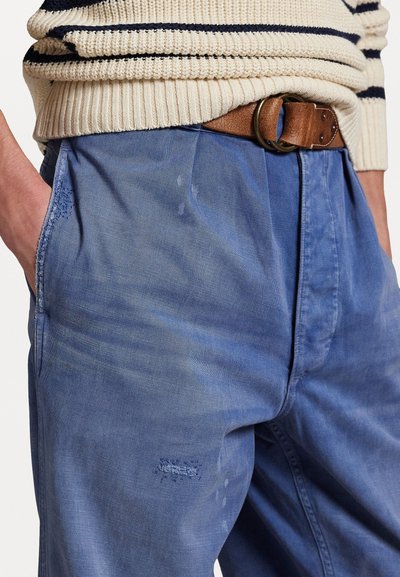 Blaue Baumwollhose mit einer lockeren Passform, ausgestattet mit seitlichen Taschen, einer verblassten Textur und einem kleinen, abgenutzten Detail nahe dem Saum. Brauner Ledergürtel.