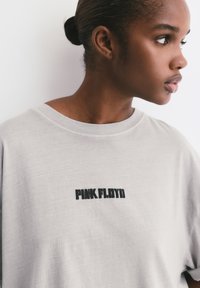 Młoda osoba z związanymi włosami, nosząca jasnoszaryT-shirt z napisem "PINK FLOYD" wydrukowanym na czarno na piersi, patrzy w prawo.