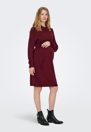 Femme enceinte en robe tricotée bordeaux à manches longues et bottines noires, debout, une main sur le ventre, sur un fond uni.