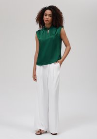 Top senza maniche in raso verde con dettaglio a chiave, abbinato a pantaloni bianchi a gamba larga; presenta una texture liscia e una vestibilità rilassata.