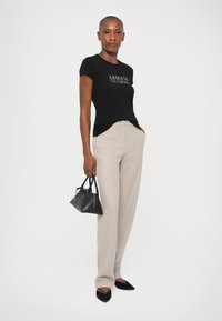 Tricou negru cu mânecă scurtă, având un logo auriu, asortat cu pantaloni largi gri deschis. Modelul ține o geantă mică neagră și poartă pantofi negri.