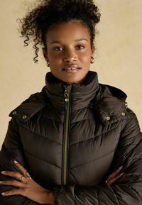 Tmavohnedá puffová bunda s vysokým golierom, horizontálnym prešívaním, zlatým zipsom a patentkami na pleciach.