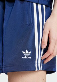 Námořnické modré sportovní šortky s bílým trojpruhovým designem na bocích. Na lemu se nachází bílé logo Adidas. Hladký materiál.