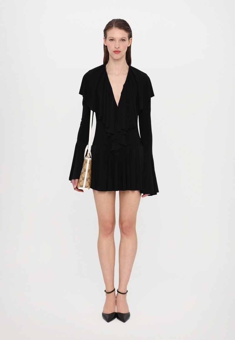 Zwarte jurk met een diepe V-hals en gekrulde details. Heeft lange mouwen en een uitlopende zoom. Gecombineerd met een gestileerde tas en zwarte hakken.