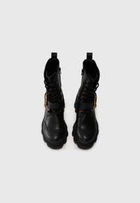 Black leather boots with a lace-up design, gold eyelets, side zipper, and chunky rubber soles. Includes a decorative golden hardware accent.