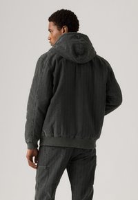 Men's dark green hooded jacket with textured fabric, ribbed cuffs and hem, featuring visible stitching and a relaxed fit.