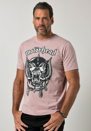 Mand med mørkt hår og skæg iført en pink Motörhead band t-shirt med en stor hærgende kraniegrafik og sorte bukser.