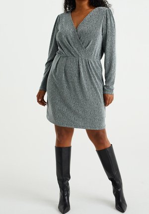 Femme portant une robe portefeuille texturée grise à manches longues, associée à des bottes noires jusqu'aux genoux, debout devant un fond uni.