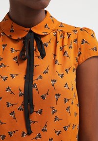 Blusa arancione a maniche corte con stampa floreale nera, colletto abbottonato e un dettaglio con cravatta nera al collo. Tessuto morbido e leggero.