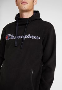 Felpa nera in pile con logo "Champion" ricamato, dotata di cappuccio con cordino e tasca frontale con zip. Texture liscia, design casual.