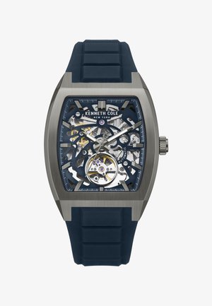 Vierkante Kenneth Cole New York automaat horloge in zilverkleurige toon met blauwe skeletwijzerplaat en zwarte rubberen band met textuur.