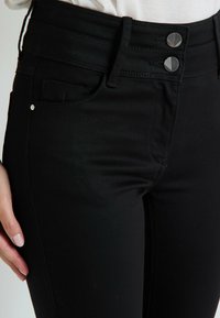 Jeans de denim negro con una cintura alta, cierre de dos botones plateados y un bolsillo delantero. La textura de la tela es suave con un ligero estiramiento.