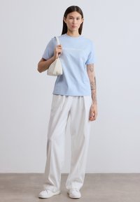 Ανοιχτό μπλε βαμβακερό μπλουζάκι με το κείμενο «Calvin Klein Jeans» τυπωμένο σε λευκό, συνδυασμένο με λευκό παντελόνι φαρδιάς γραμμής και μικρή κρεμ τσάντα.