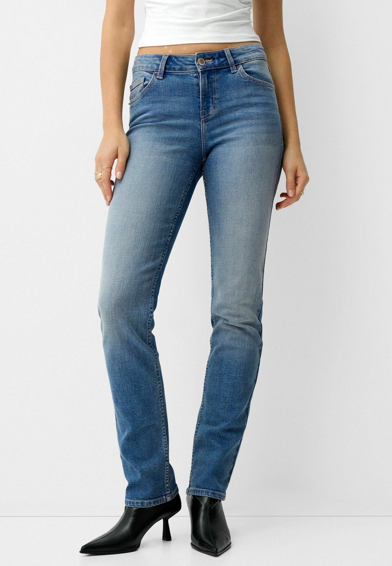 Femme portant un jean slim bleu clair, un top court blanc et des bottines noires à talons et bout pointu sur fond uni.