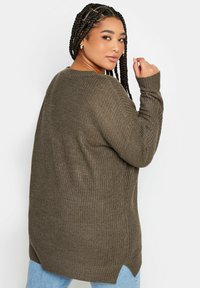 Olivgrüner Strickpullover mit lockerer Passform, gerippter Textur, seitlichen Schlitzen und langen Ärmeln, kombiniert mit hellblauen Jeans.