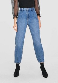 Vaqueros de mezclilla azul de cintura alta con pierna recta, que presentan bolsillos frontales y aberturas laterales, combinados con botas negras hasta el tobillo y una blusa negra.