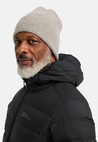 Giacca puffa nera con colletto alto, caratterizzata da un logo. Berretto a coste grigio. Barba corta e capelli rasati. Profilo laterale, sfondo neutro.