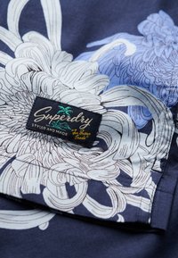 Tejido azul marino con patrón floral blanco; presenta una etiqueta negra con "Superdry" y el logo de una palmera; textura suave, diseño vibrante.