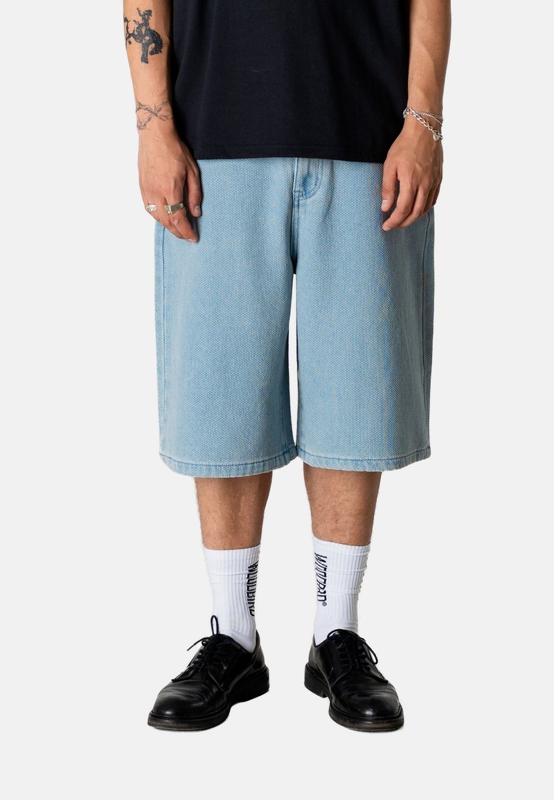 Homme portant un short en jean bleu clair jusqu'aux genoux, des chaussettes blanches avec du texte noir, des chaussures en cuir noires et des bijoux en argent aux poignets et aux doigts.