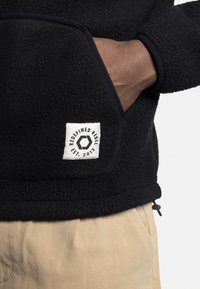 Pull noir en polaire avec une surface texturée, dotée d'une poche avant et d'une étiquette tissée portant "REDEFINED REBEL". Cordon de serrage à l'ourlet.