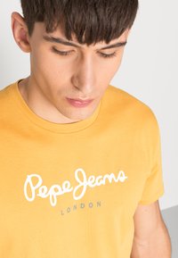 Geltona medvilninė marškinėliai su baltu "Pepe Jeans" logotipu ir "LONDON" pilka spalva, apvaliu iškirpimu ir trumpomis rankovėmis.