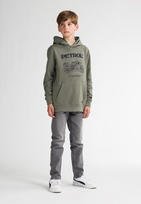 Sudadera verde oliva con un estampado gráfico y bolsillo frontal, combinada con jeans grises y zapatillas blancas con detalles negros.