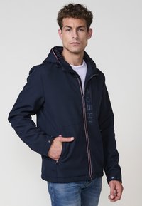 Chaqueta con capucha azul marino, con cremallera frontal completa, dos bolsillos frontales y detalles en rojo y blanco en la cremallera y los puños.