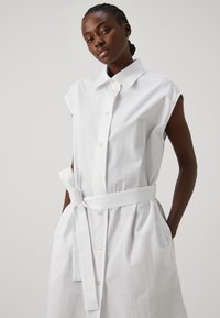Abito camicia bianco a maniche corte con tessuto testurizzato, chiusura frontale a bottoni e cintura da annodare in vita, dotato di tasche laterali.