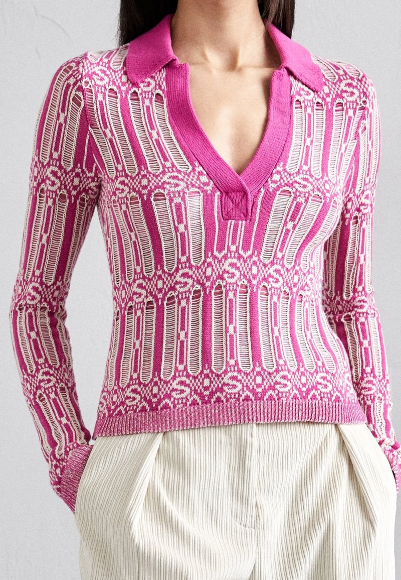 Femme portant un pull en tricot rose et blanc à motifs avec un col et un profond décolleté en V, associé à un pantalon en velours côtelé crème taille haute.