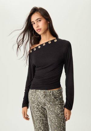 Young woman with long dark hair wears black off-shoulder top with silver eyelets and leopard-print pants against a plain light background.