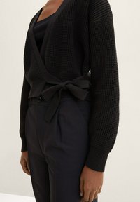 Černý pletený kardigan s konstrukcí do zavinovacího stylu, s dlouhými rukávy a zavazovacím pasem. V kombinaci s černými kalhotami s vysokým pasem.