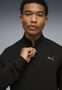 Sudadera negra con cremallera y cuello alto, que presenta un pequeño logo de Puma gris en el pecho. Textura suave y diseño ajustado.