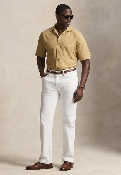Mann trägt beige kurzärmliges Hemd mit Knöpfen, weiße Hose, braunen Gürtel, braune Schuhe, Sonnenbrille und Uhr, steht mit einer Hand in der Tasche.