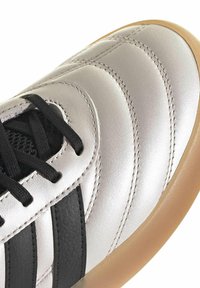 Zapatilla blanca con rayas negras, cordones negros, costuras detalladas y suela de goma color caramelo, mostrada en primer plano desde un ángulo superior frontal.