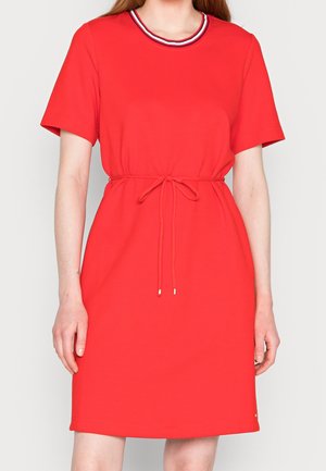 Kobieta ubrana w czerwoną sukienkę z krótkim rękawem, z talią ze sznurkiem oraz okrągłym dekoltem wykończonym białym i granatowym paskiem.