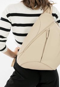 Beige slingväska med en elegant design, tillverkad av ett glatt material. Har ett vinklat zipfack och justerbart band, bärs över ryggen.