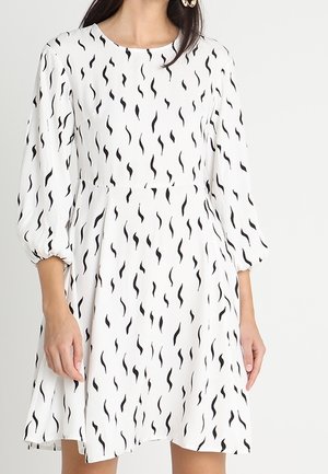 Femme portant une robe blanche ornée de motifs abstraits noirs en forme de zigzags verticaux et de manches bouffantes trois-quarts.