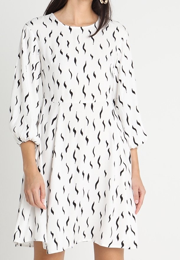 Femme portant une robe blanche ornée de motifs abstraits noirs en forme de zigzags verticaux et de manches bouffantes trois-quarts.