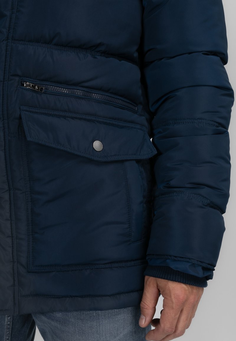 Veste matelassée bleu marine avec une poche zippée et une poche à rabat à fermeture à pression, dotée de poignets en côte et d'une texture lisse.