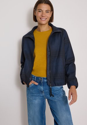 Femme souriante portant une chemise jaune moutarde, une veste zippée bleu marine et un jean bleu, debout la main dans la poche devant un fond uni.