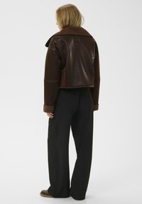 Donna di spalle che indossa una giacca in pelle marrone corta con colletto e polsini in shearling, pantaloni neri a gamba larga e scarpe marroni.